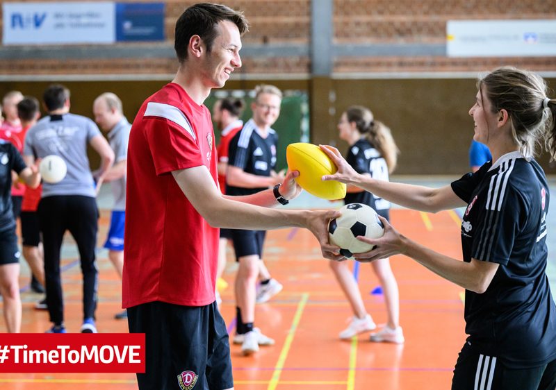 Im Vordergrund stehen ein Mann und eine Frau in Sport-Klamotten. Sie übergeben sich zwei verschiedene Bälle. Im Hintergrund sind weitere Personen in Bewegung zu sehen.