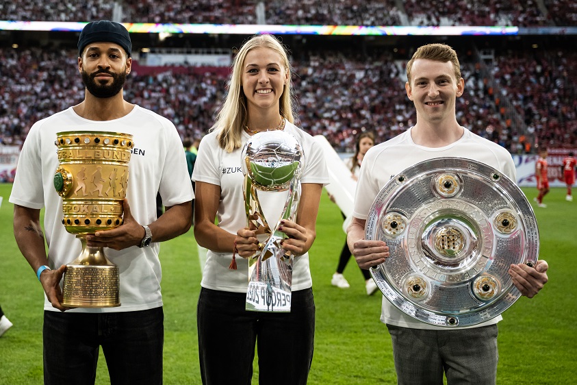 Eine Athletin und zwei Athleten halten den DFB-Pokal, den Supercup und die Meisterschale hoch.