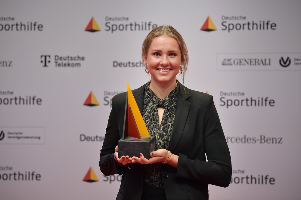Lisa Spark steht vor einer Pressewand mit verschiedenen Logos und hält eine Trophäe in den Händen.