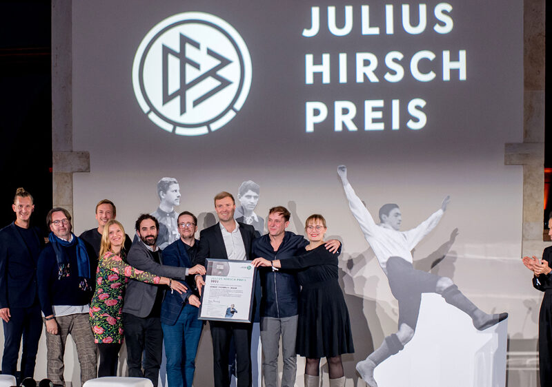 Auf einer Bühne stehen neun Personen. Fünf von ihnen halten eine Urkunde in der Hand. Im Hintergrund steht an die Wand projiziert: Julius Hirsch Preis.