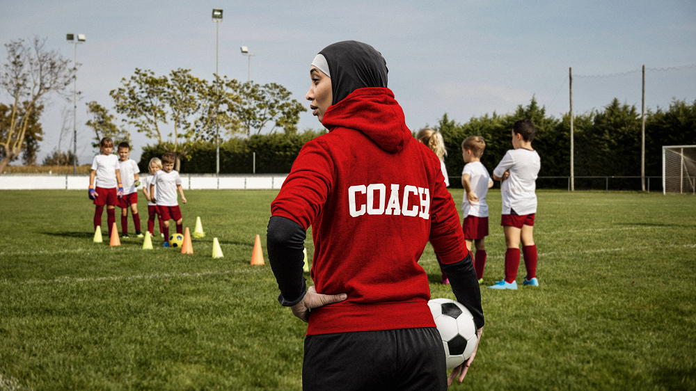 Eine Trainerin hält einen Ball in den Händen und steht auf dem Trainingsplatz, während Kinder eine Fußballübung durchführen.