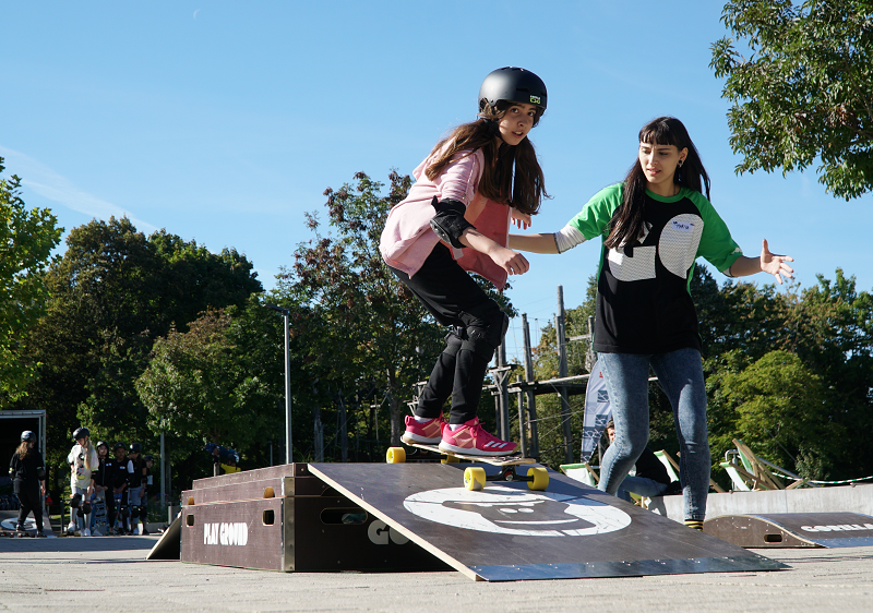 Ein Mädchen übt das Skateboardfahren, eine Betreuerin hilft ihr dabei.