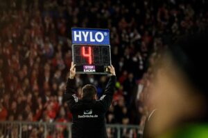 Eine Person hält eine elektronische Auswechseltafel mit der Aufschrift "STOP RACISM" im Fußballstadion hoch. Die Anzeigetafel zeigt die Zahl 42. Im Hintergrund sind verschwommene Zuschauerreihen zu sehen.