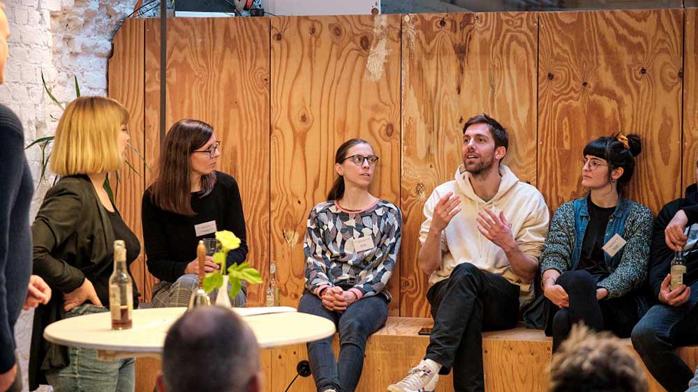 Gruppe von Menschen sitzt in einem Workshop-Setting auf einer Holzbank und diskutiert. Ein Mann gestikuliert dabei lebhaft, während die anderen ihm aufmerksam zuhören. Im Vordergrund steht ein Tisch mit Wasserflaschen und Blumen.