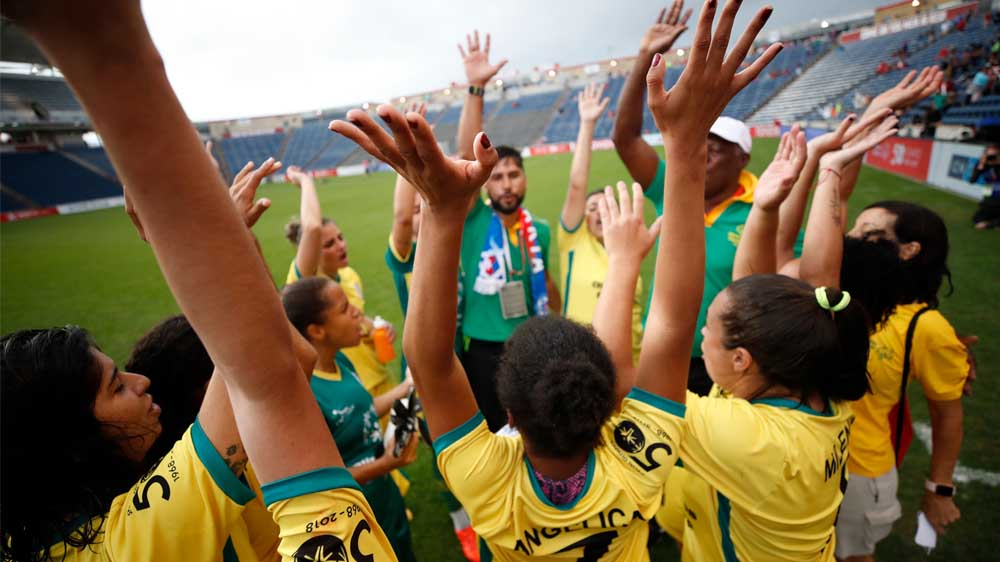 Eine Gruppe junger Menschen steht auf einem Fußballfeld im Kreis. Die jungen Menschen heben ihre Arme in die Luft.