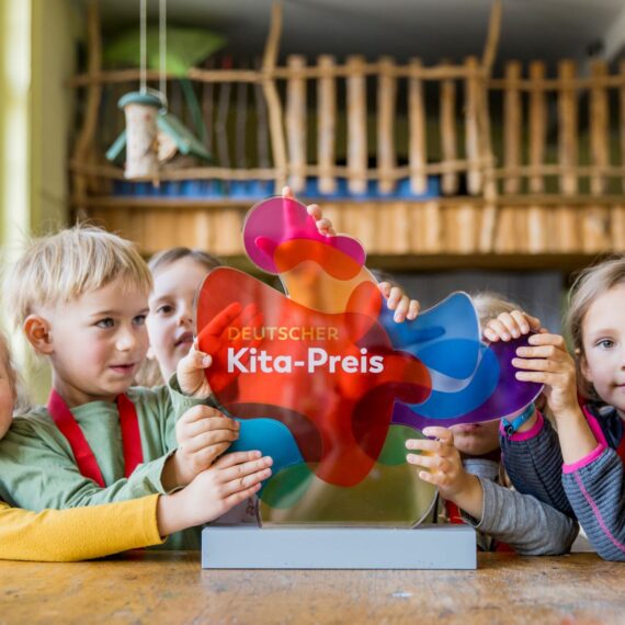 Eine Gruppe von Kindern steht erwartungsvoll vor einer farbenfrohen Trophäe mit der Aufschrift "Deutscher Kita-Preis".