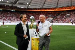 Joel Beckenbauer steht neben seinem Vater, der Fußballlegende Franz Beckenbauer, vor der MHPArena in Stuttgart. Beide werden interviewt, während im Hintergrund der Supercup-Pokal zu sehen ist.