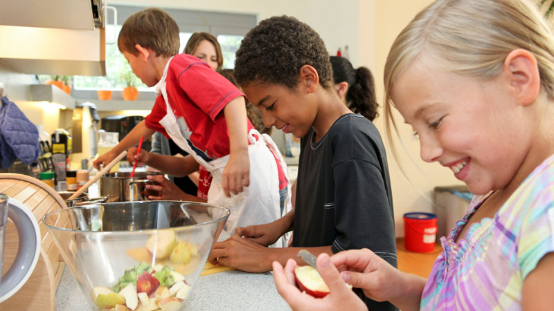 Kinder unterschiedlichen Geschlechts und unterschiedlicher Hautfarbe lächeln gemeinsam und schneiden dabei Obst in kleine Stücke, um einen Obstsalat in einer Schüssel zuzubereiten.