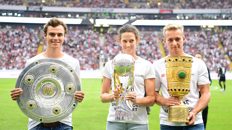 Die Sportler Vinzenz Geiger, Lena Schöneborn und Johannes Floors stehen in einem Fußballstadion vor dem Anpfiff des Supercups. Jeder von ihnen hält einen Pokal in den Händen.