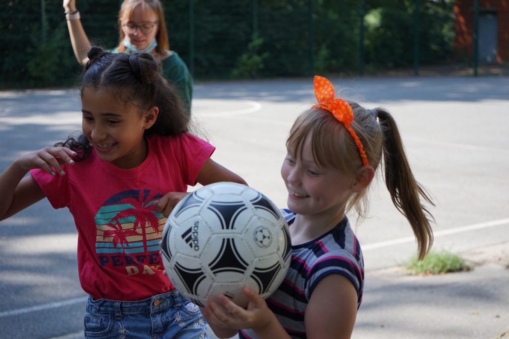 Zwei Mädchen lächeln im Vordergrund des Bildes. Das eine Mädchen hält einen Fußball in den Händen.