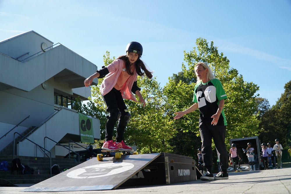 Ein Mädchen in rosa T-Shirt und Jacke und einer schwarzen Sporthose fährt mit dem Skateboard eine Rampe herunter. Sie wird dabei von einer Workshop-Leiterin beobachtet.